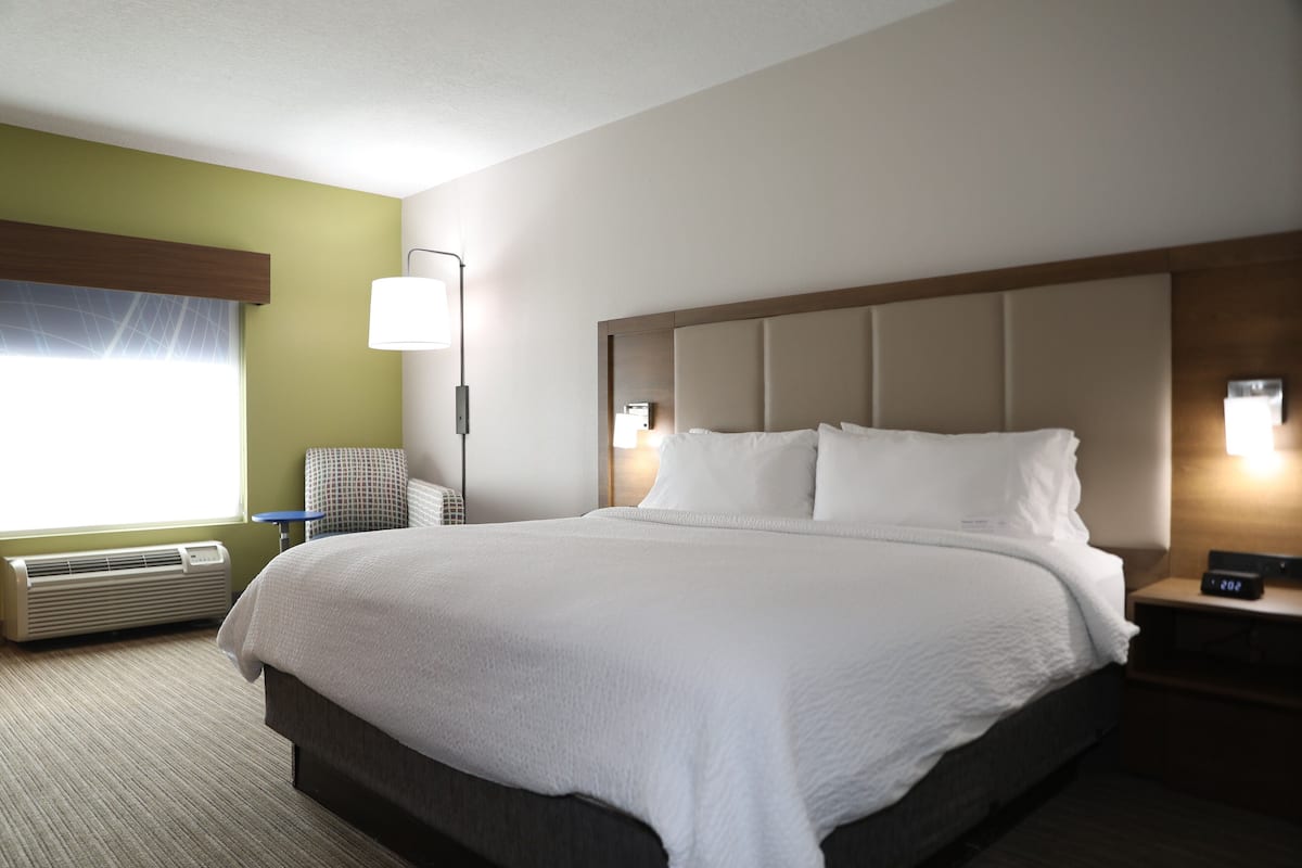 A hotel room with a large bed, a bedside table with a lamp, a chair, and a window with curtains.