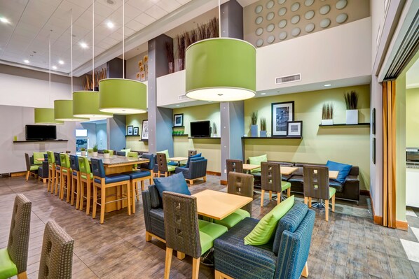 A modern dining area with multiple tables and chairs, green and blue seating, and pendant lighting. - Hampton Inn & Suites Sarasota/Bradenton-Airport, FL (Sarasota)