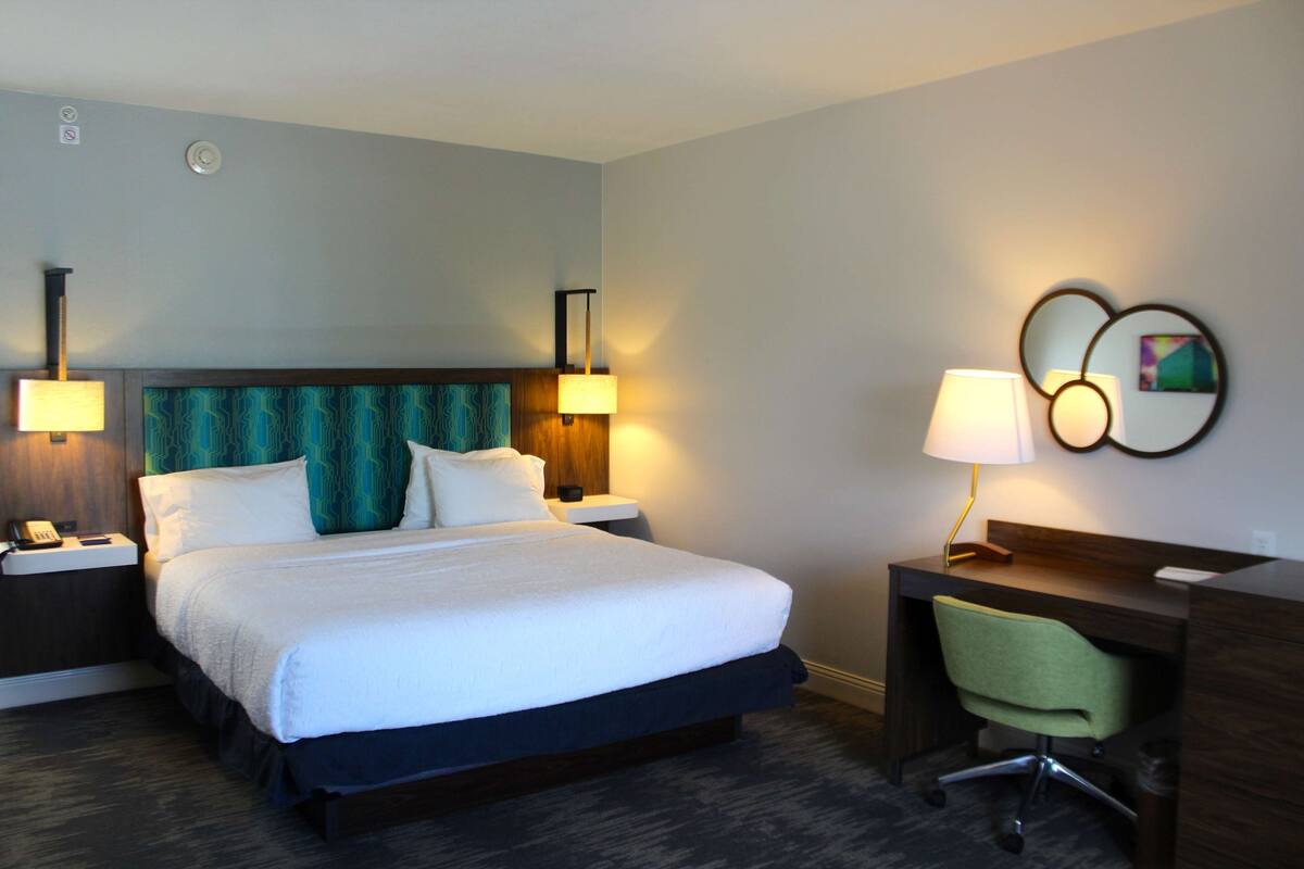 A hotel room with a large bed, two bedside tables with lamps, a desk with a chair, and a wall-mounted artwork.