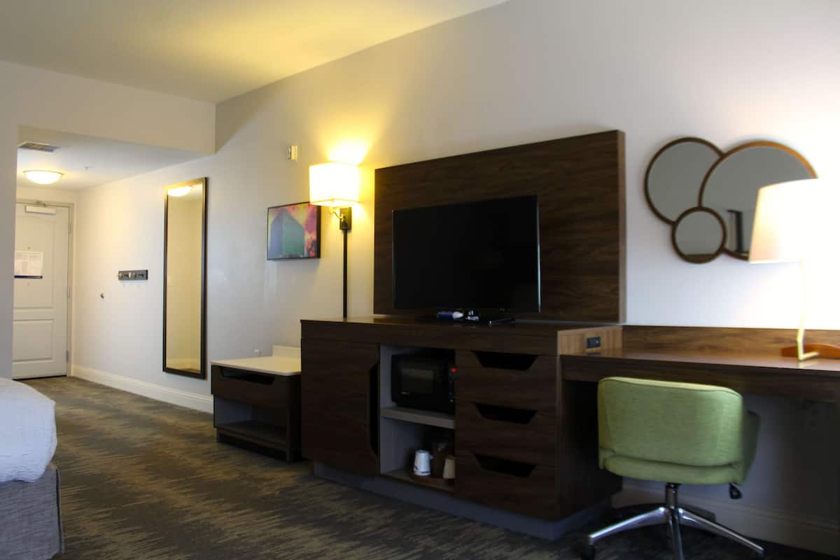 A hotel room with a TV, a desk with drawers, a green chair, a microwave, and a large mirror.
