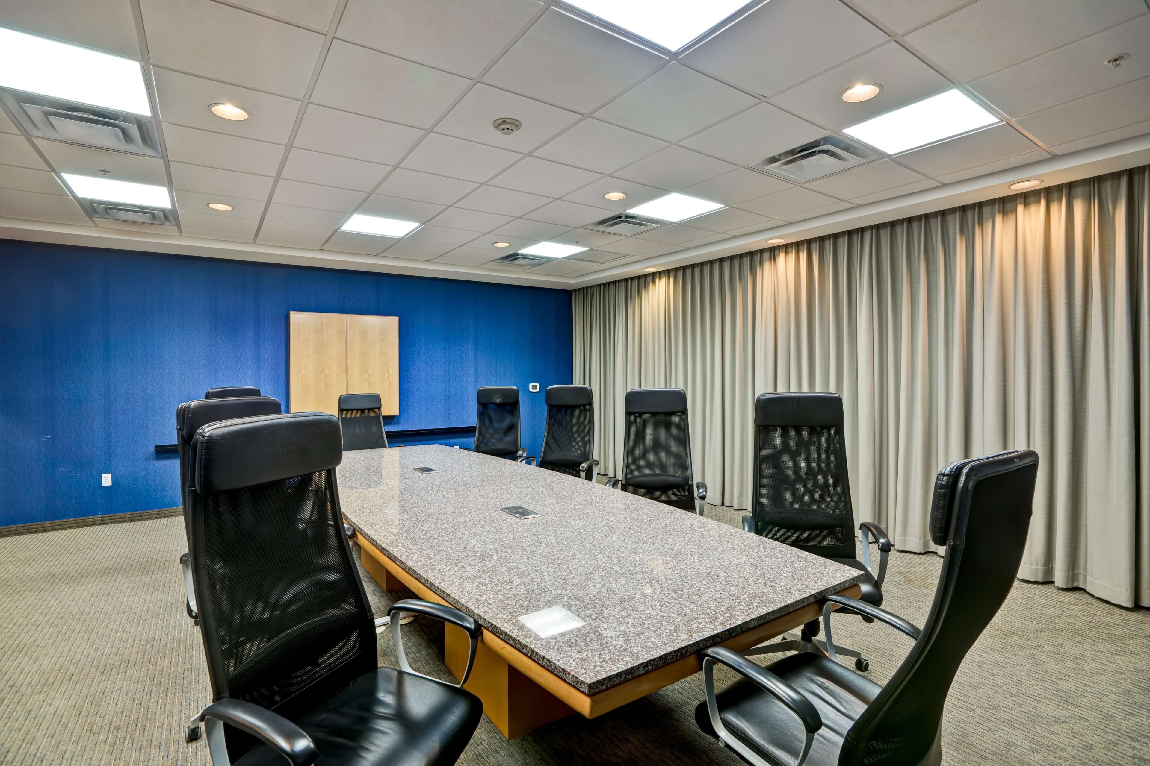 A conference room with a long table, six black chairs, a blue accent wall, and a large window with curtains.