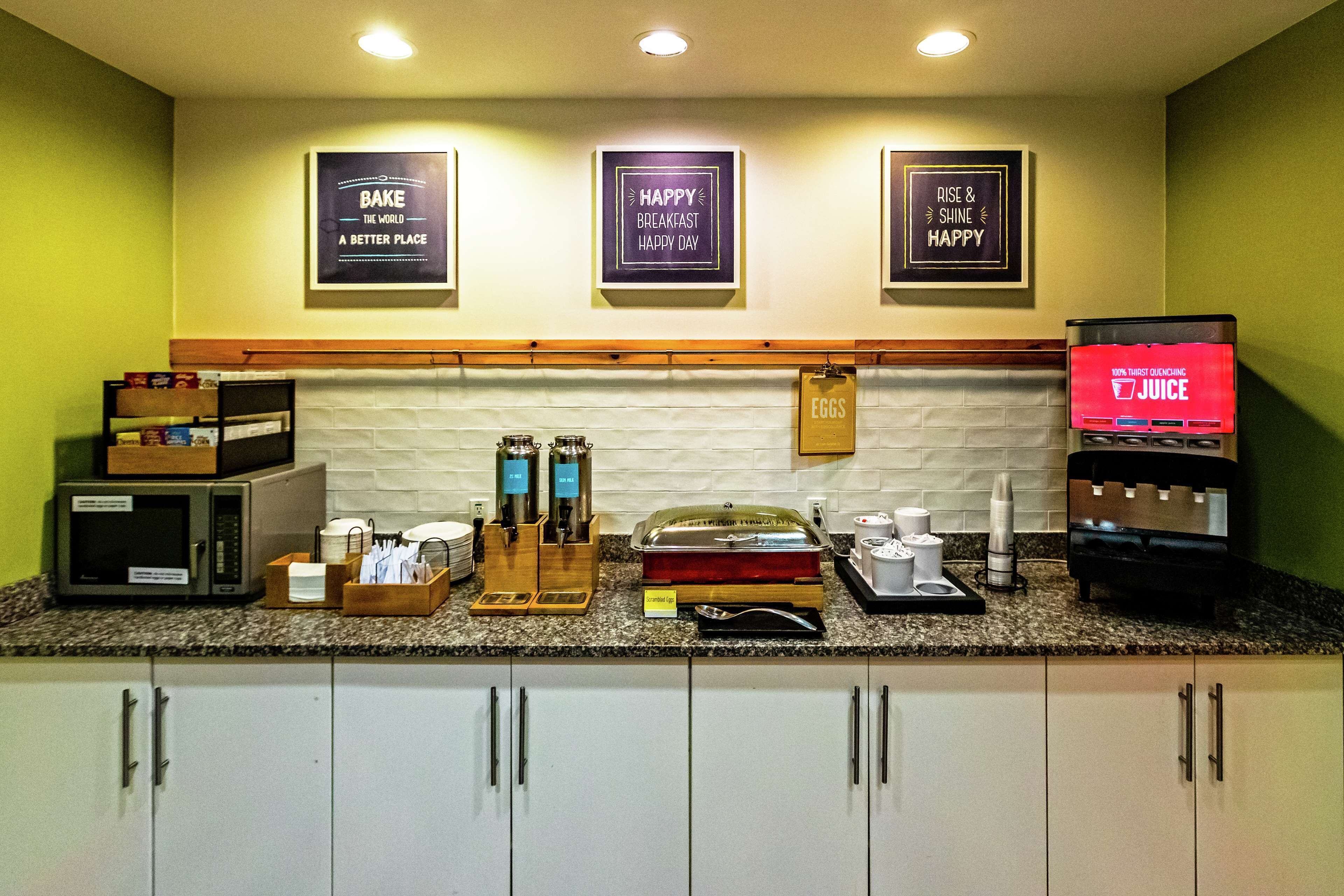 A breakfast bar with a microwave, coffee maker, and juice dispenser. There are framed signs on the wall and a shelf with food items.