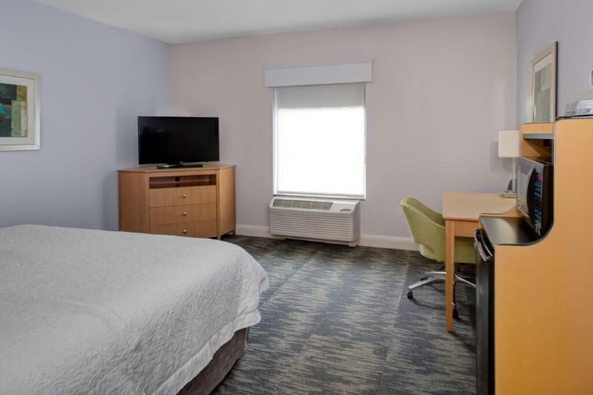 Habitación de hotel con cama, escritorio, silla, televisor y unidad de aire acondicionado.