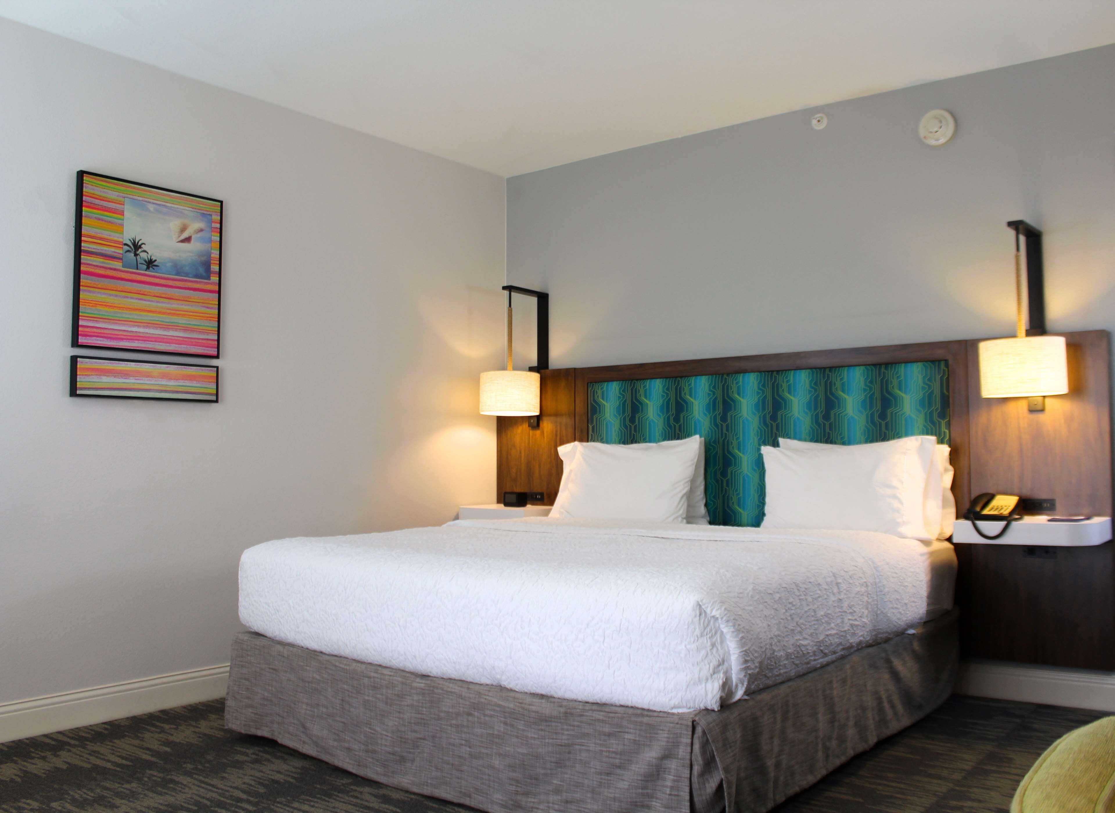 A hotel room with a large bed, two bedside lamps, a headboard with a geometric pattern, and a wall-mounted clock.
