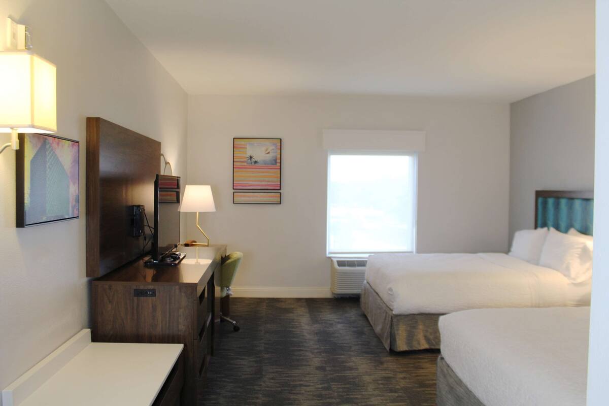 A hotel room with a desk, chair, two beds, a TV, and a window with curtains.