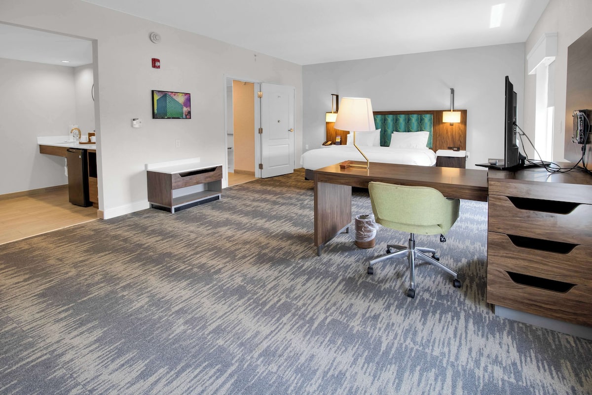 A hotel room with a bed, desk, chair, and a small kitchenette area.