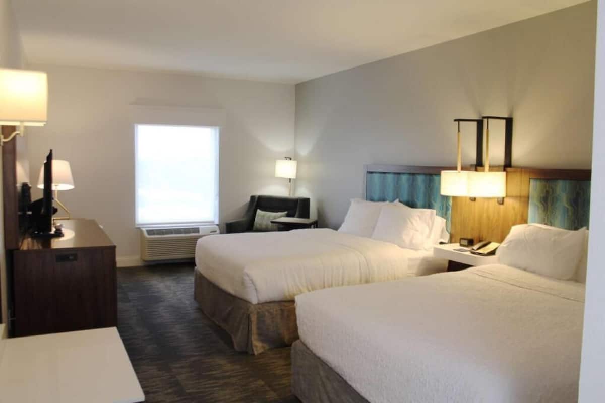 A hotel room with two beds, a desk, a chair, a lamp, and a window.