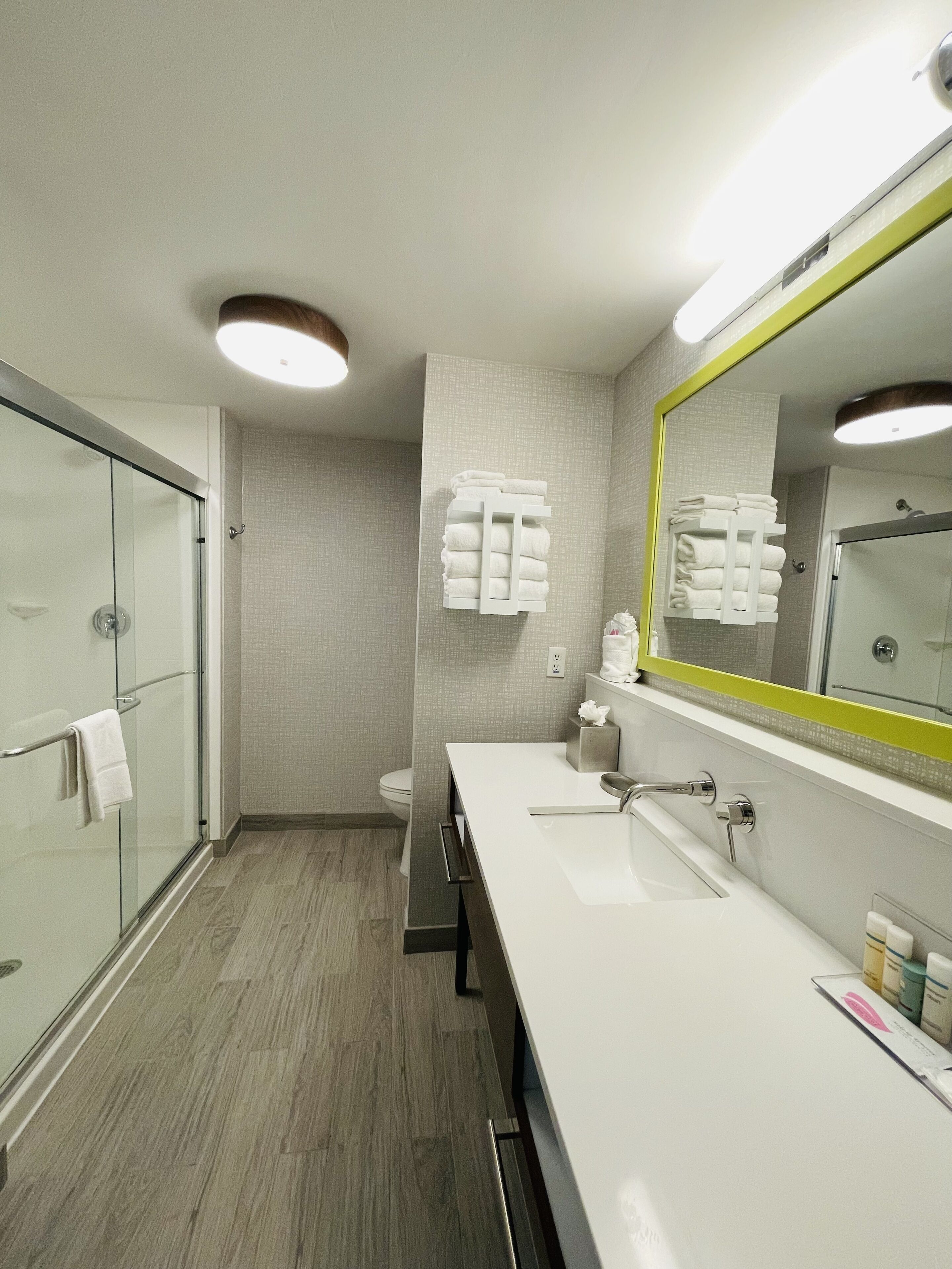 Un baño moderno con cabina de ducha de vidrio, un lavabo blanco con un espejo grande y toallas ordenadas en un escurridor de pared.