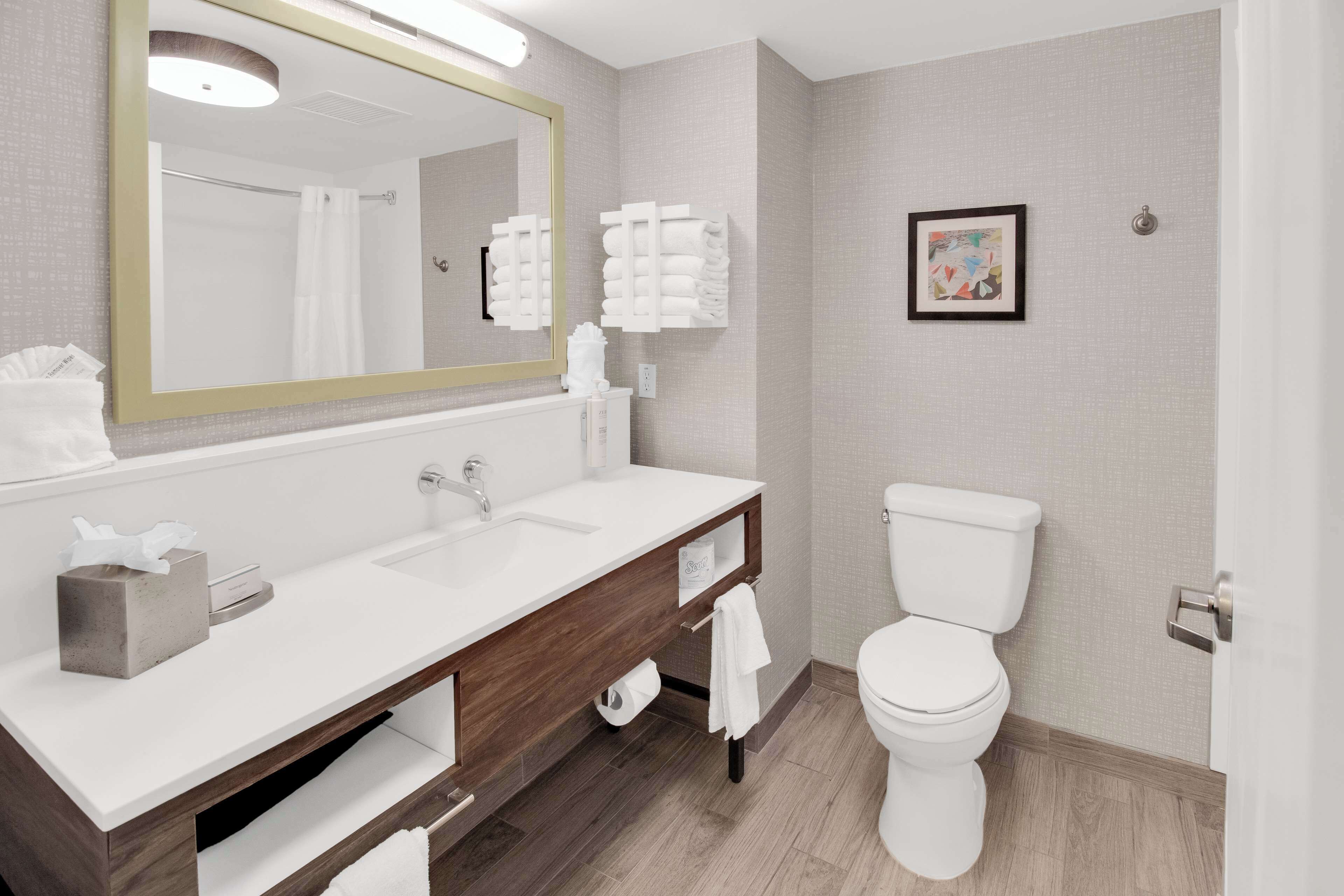A modern bathroom with a large mirror, a white sink, a wooden vanity, a toilet, and a framed picture on the wall.