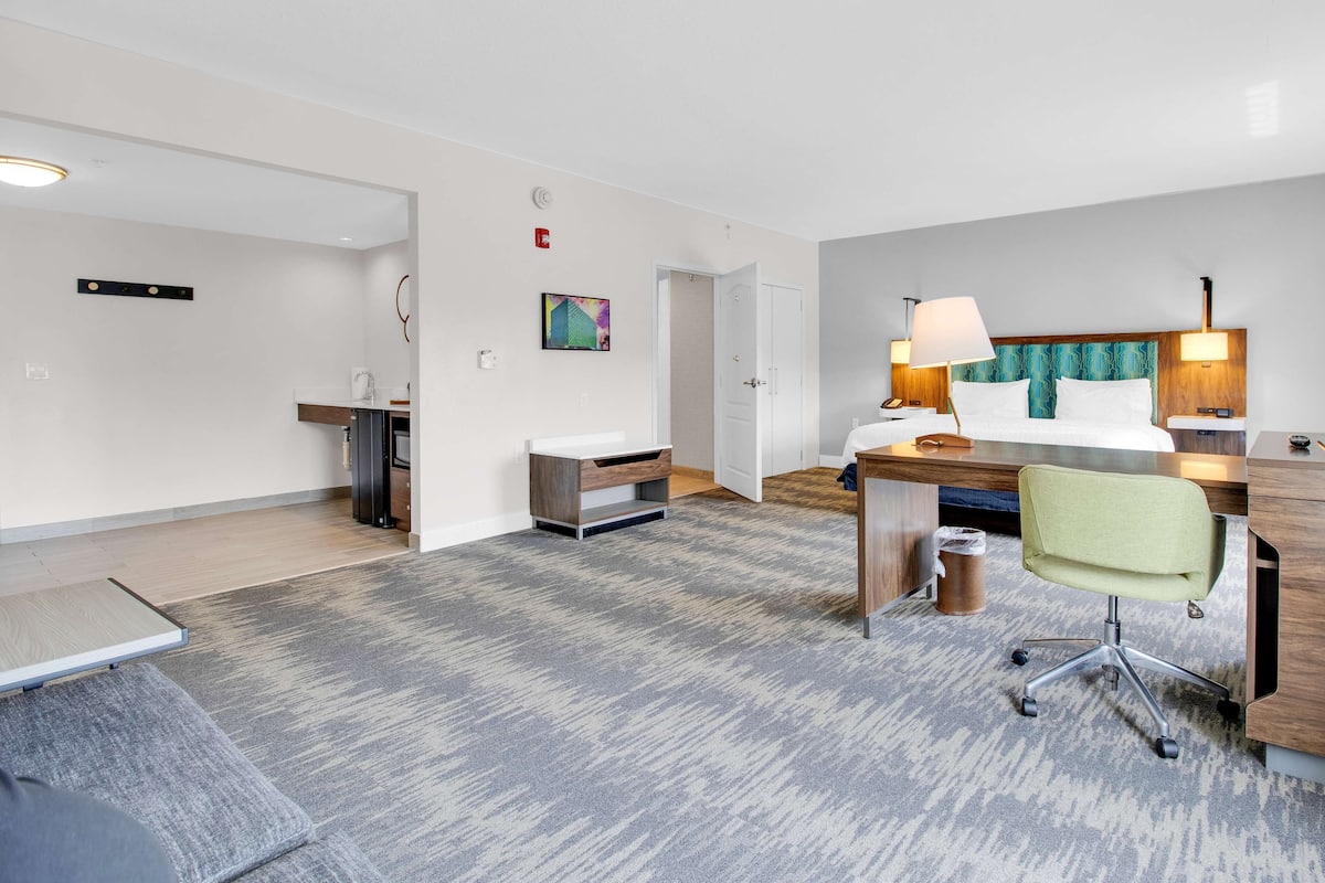 A hotel room with a bed, desk, chair, and a small kitchenette.