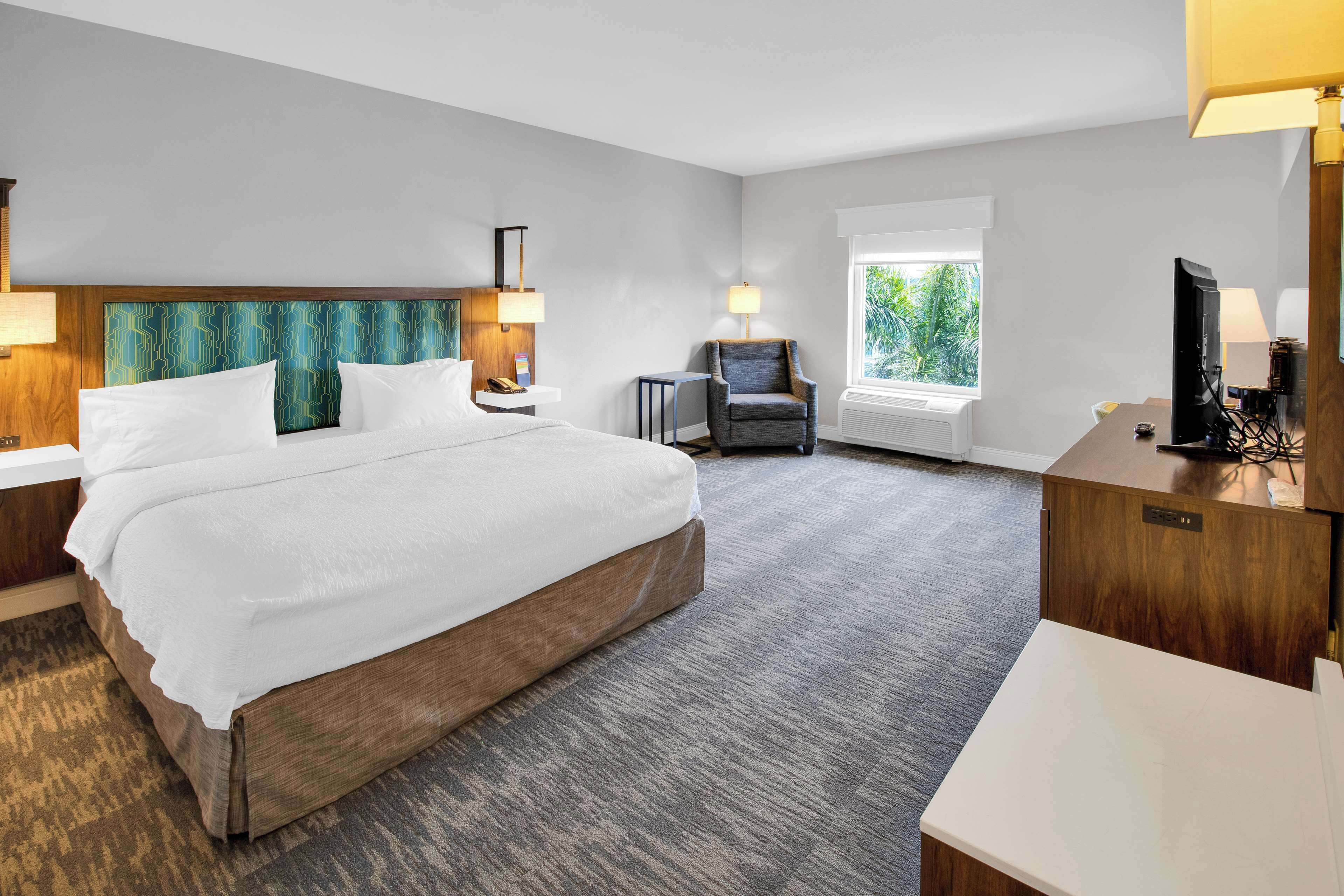Ein Hotelzimmer mit einem großen Bett, einem Schreibtisch mit Computer, einem Sessel und einem Fenster mit Blick auf Grünflächen.