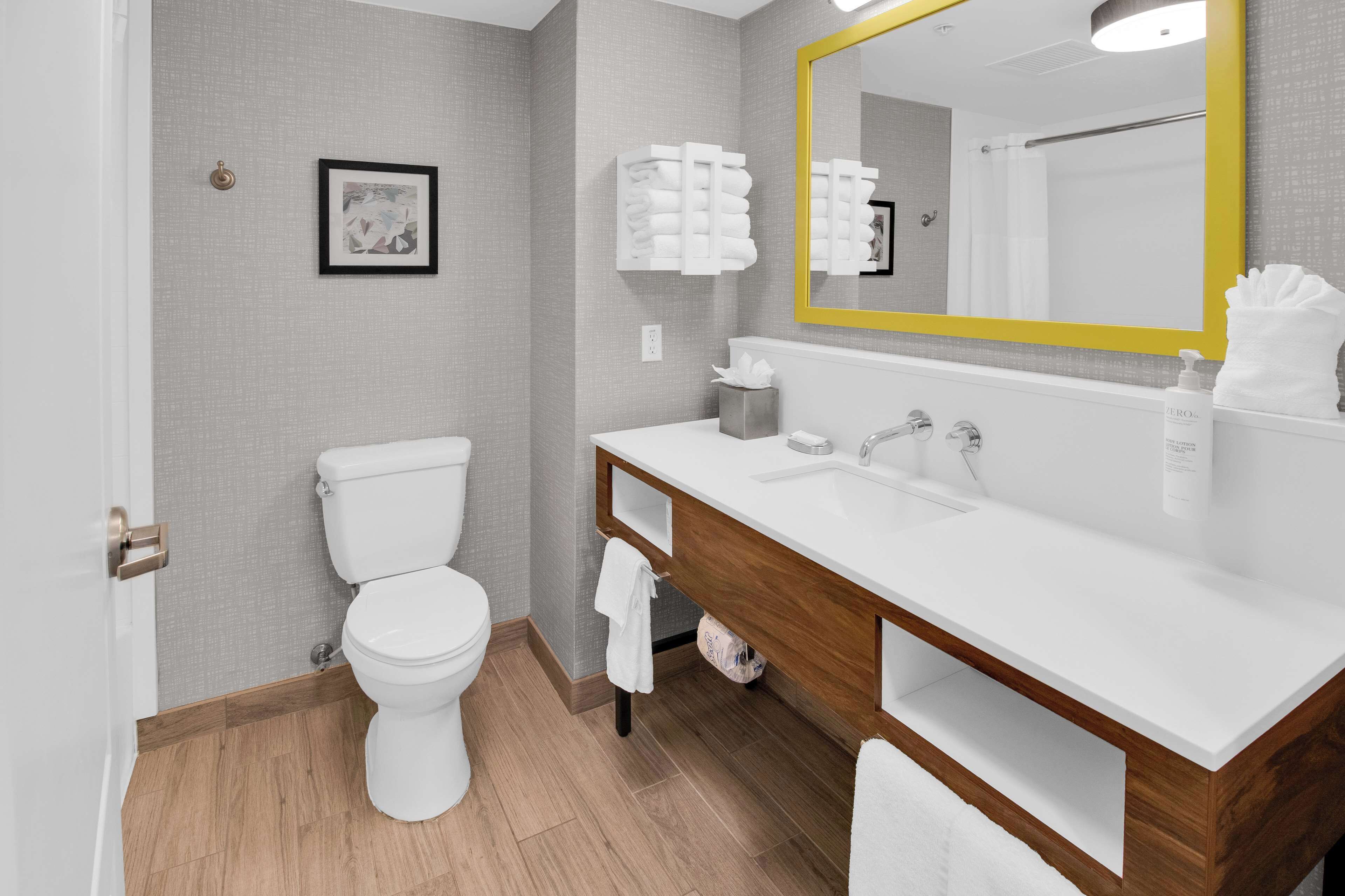 A modern bathroom with a white toilet, a wooden vanity with a white sink, and a large mirror with a yellow frame.