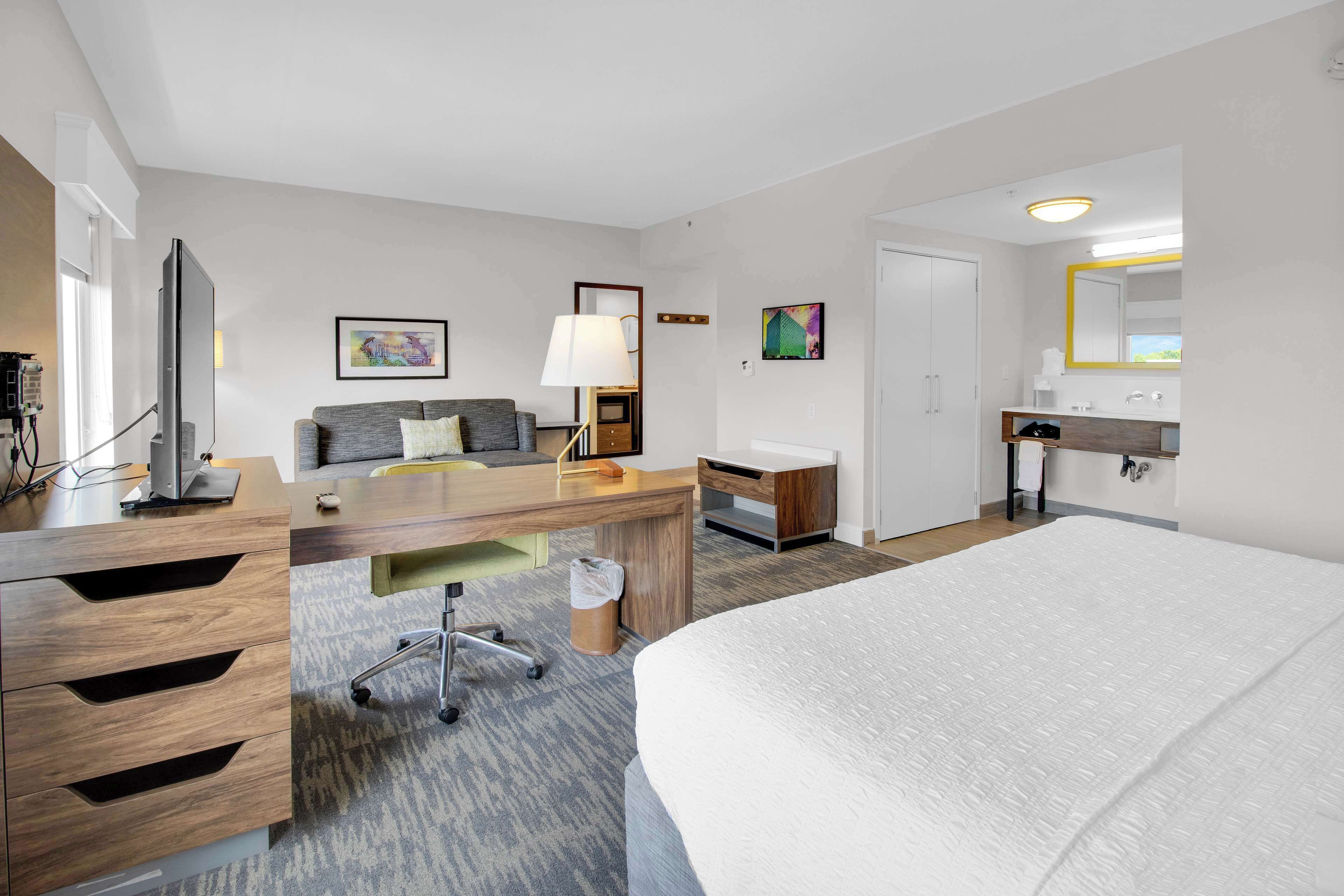 Habitación de hotel con una cama grande, un escritorio con computadora, un sofá, una lámpara y un baño con espejo y lavamanos.