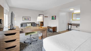Habitación de hotel con una cama grande, un escritorio con computadora, un sofá, una lámpara y un baño con espejo y lavamanos.