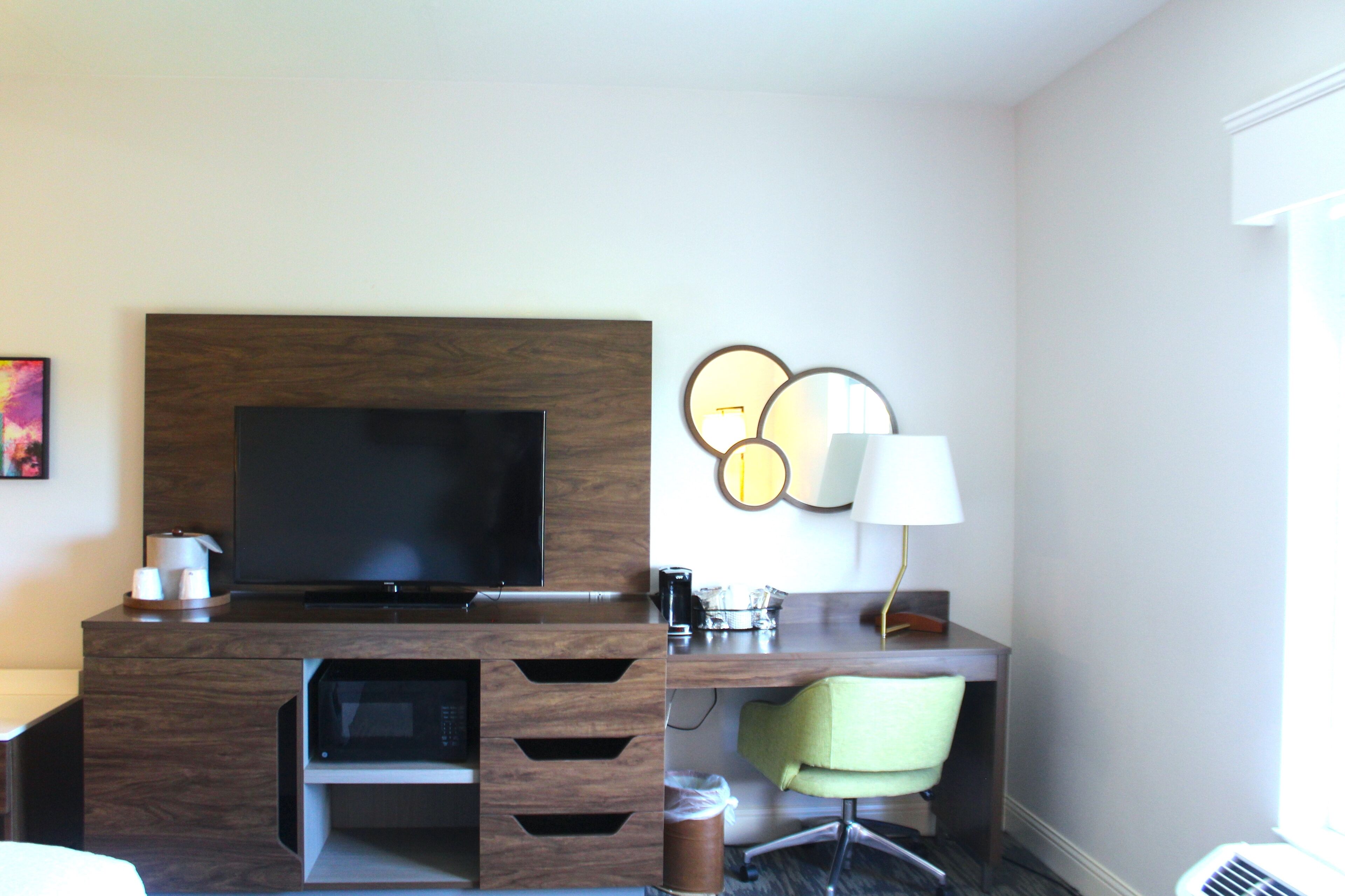 Habitación de hotel con televisor de pantalla plana, escritorio con lámpara, silla y cafetera.