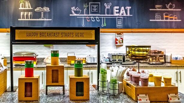 A breakfast bar with a chalkboard sign that reads "HAPPY BREAKFAST STARTS HERE" and a variety of food and drink options. - Hampton Inn & Suites Sarasota/Bradenton-Airport, FL (Sarasota)