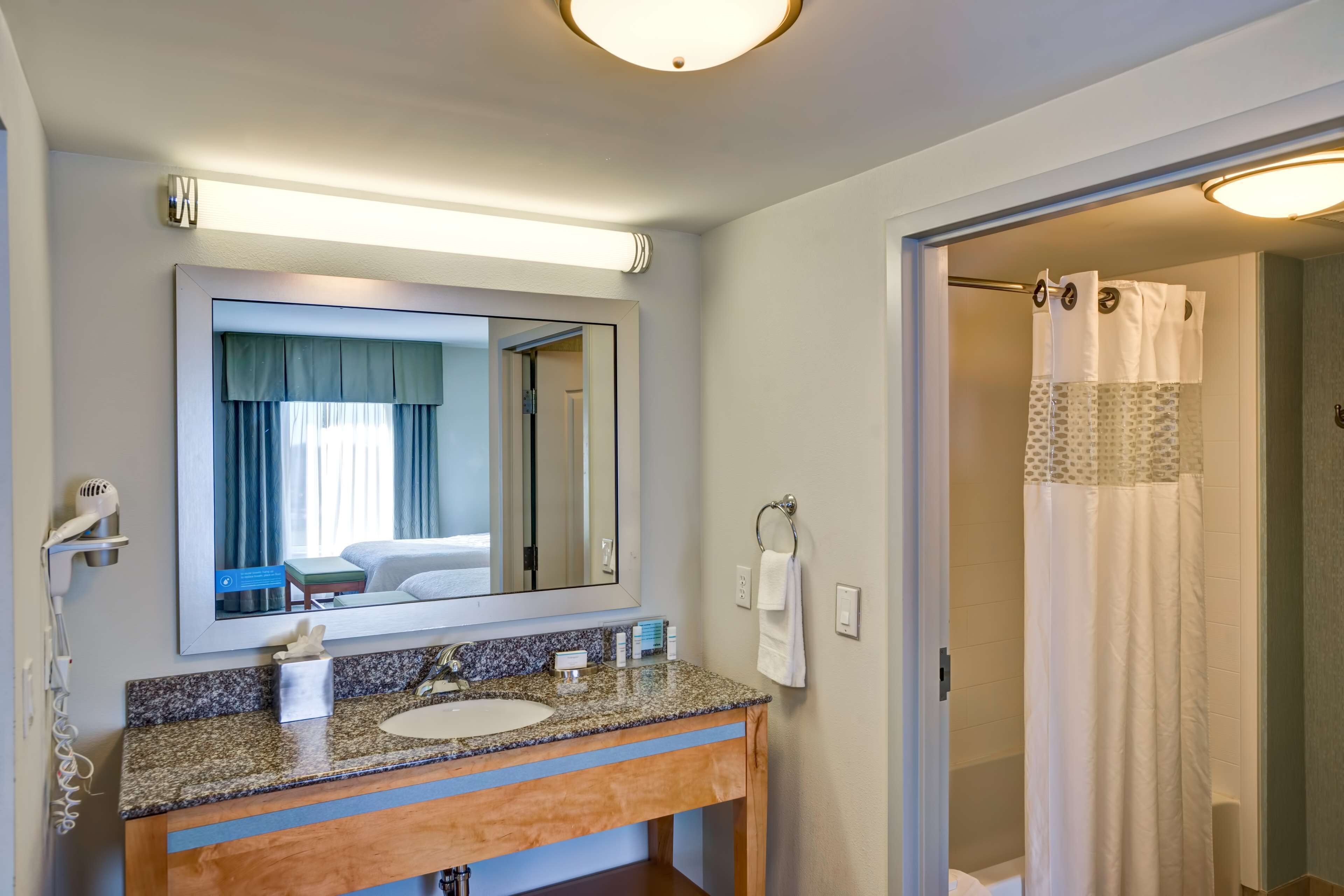 A bathroom with a granite countertop sink, a mirror, a hairdryer, and a shower with a white curtain.