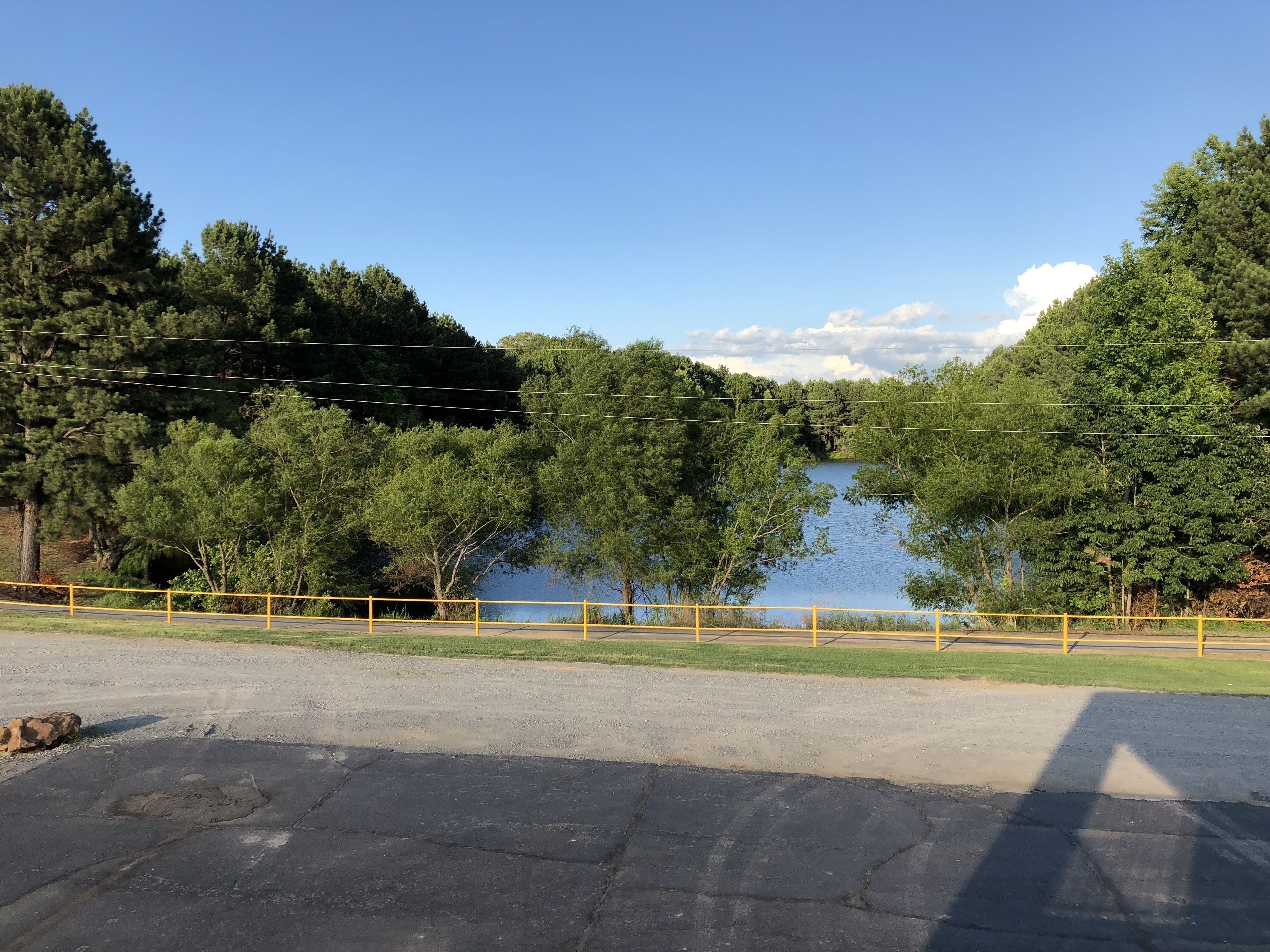 A parking lot with a yellow fence, a paved road, and a body of water surrounded by trees.