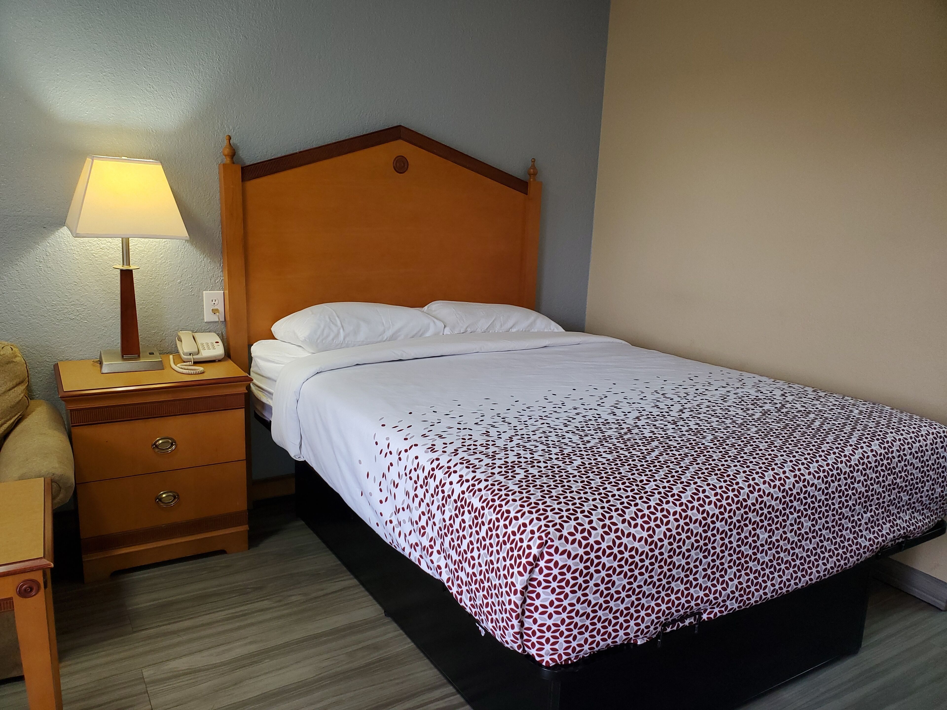 A hotel room with a wooden bed, white bedding, a bedside lamp, a telephone, and a wooden nightstand.