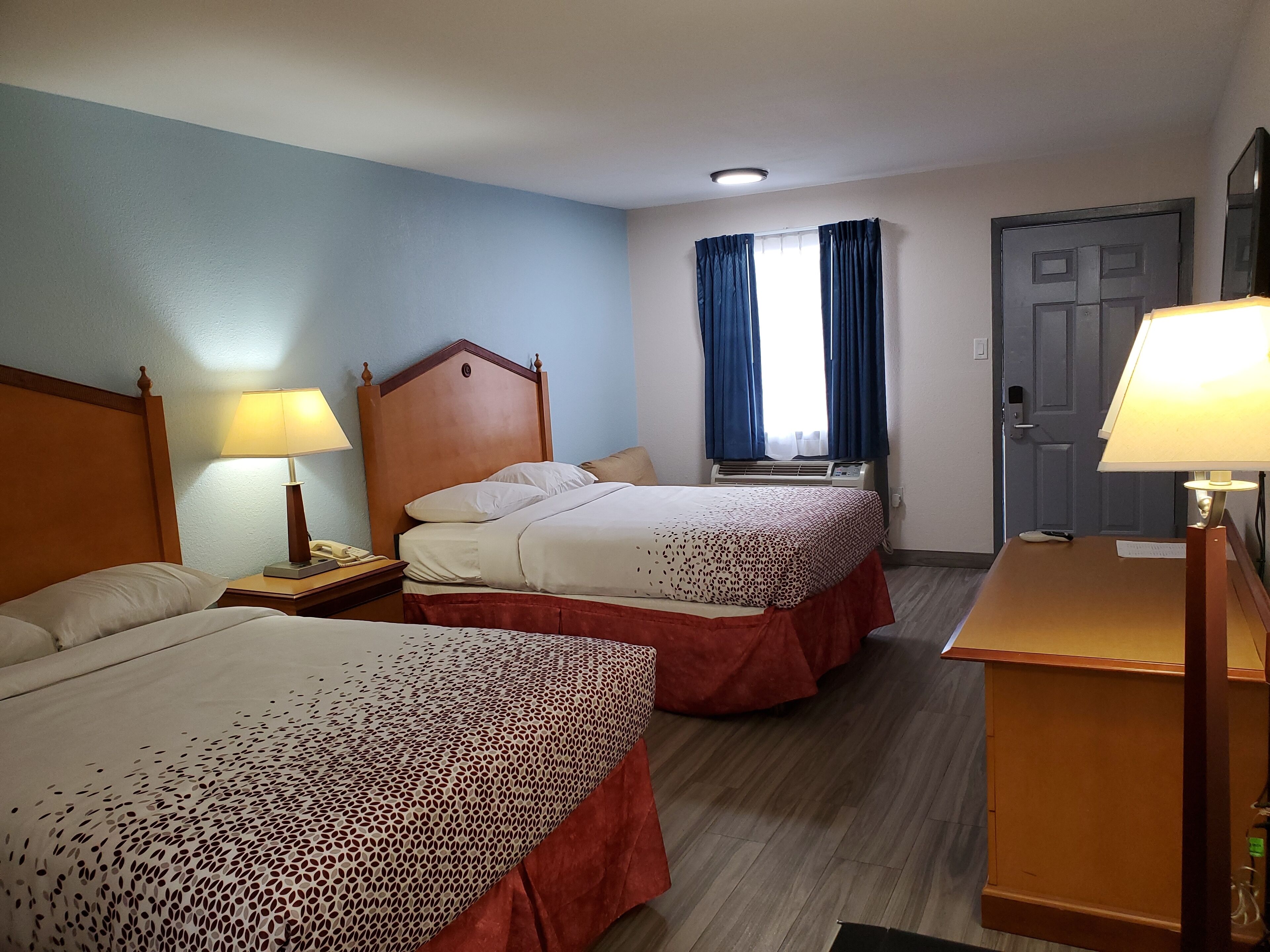 A hotel room with two beds, a desk, a lamp, a window with curtains, an air conditioning unit, and a door.