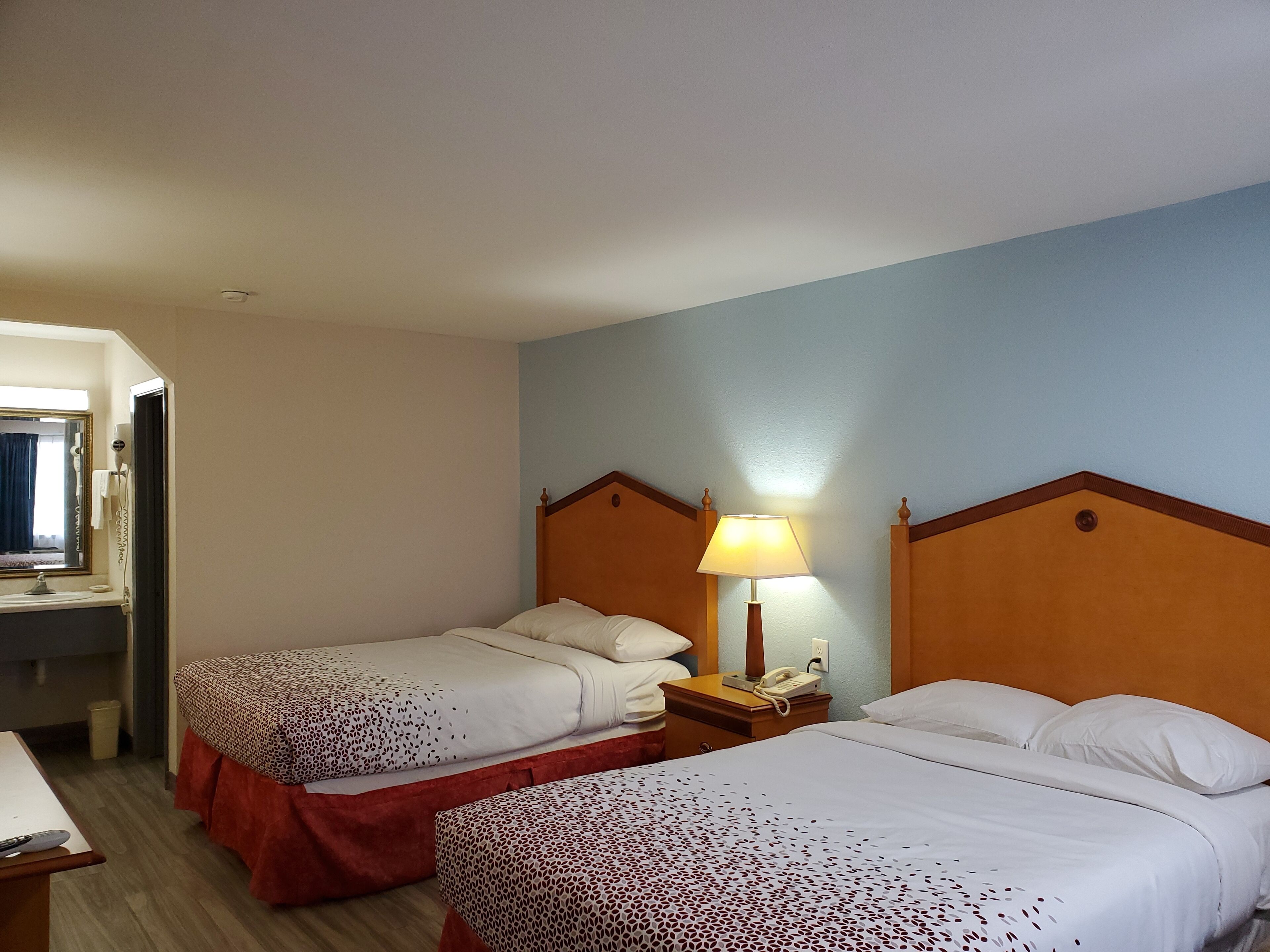 Une chambre d’hôtel avec deux lits, une table de chevet avec une lampe, un miroir et une salle de bain visible à travers une porte ouverte.