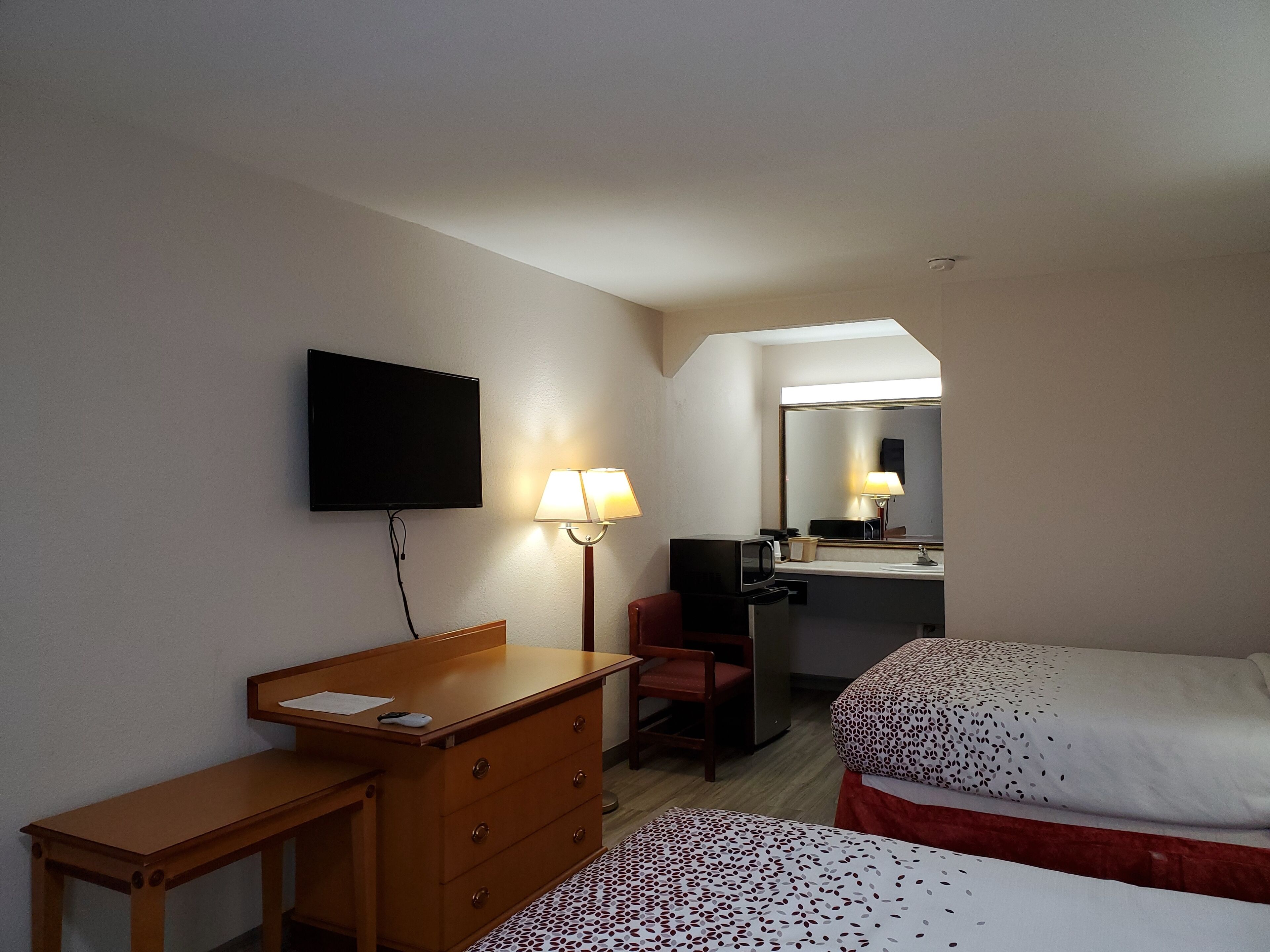 A hotel room with a wooden desk, a chair, a bed with patterned bedding, a TV mounted on the wall, and a mirror above the sink.