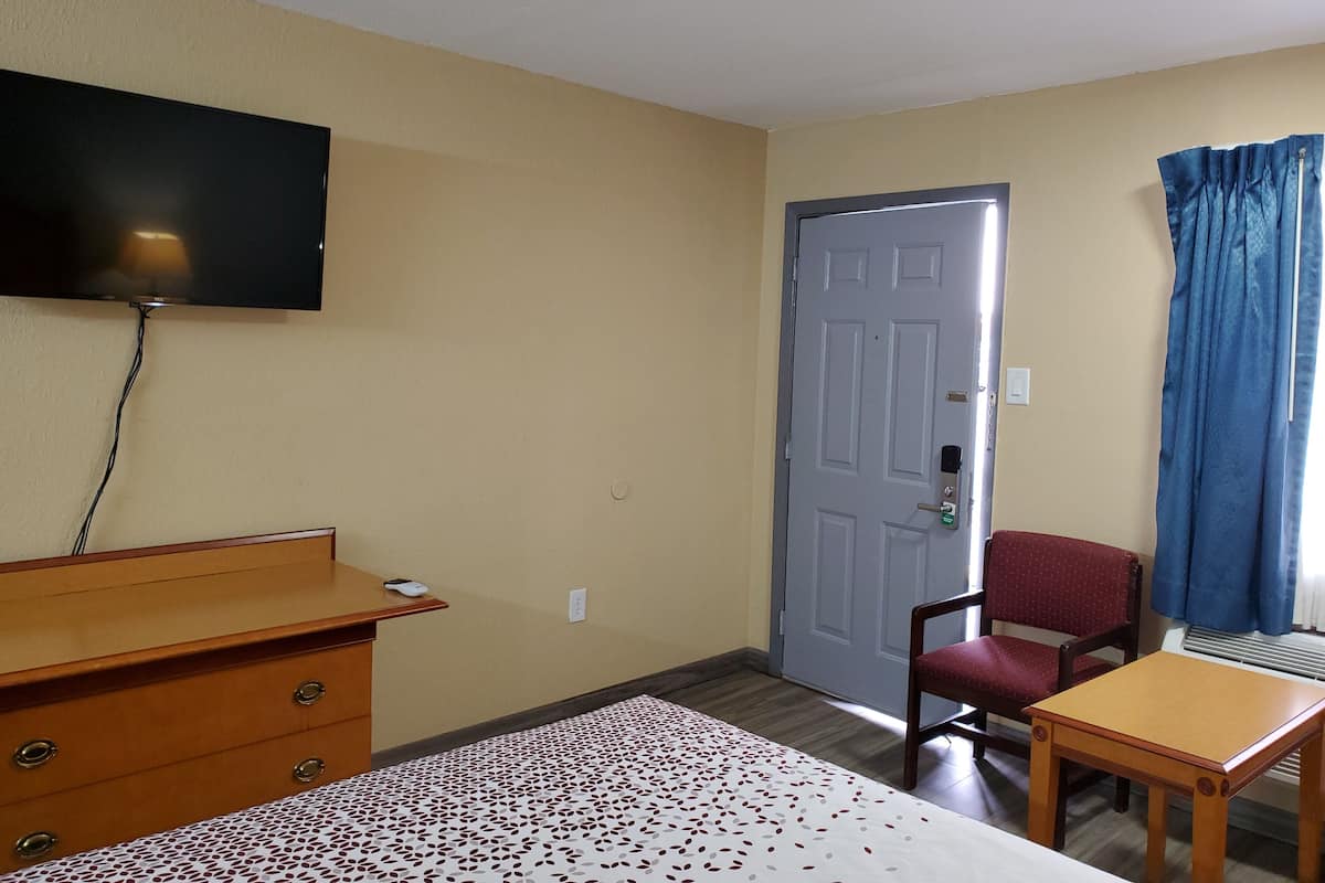 Une chambre d’hôtel comprenant un lit, une commode en bois, un bureau, une chaise, un téléviseur fixé au mur et une porte avec une poignée.