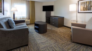 Habitación de hotel con sofá, una mesita de centro redonda, televisor de pantalla plana, escritorio con cajones, lámpara y un cuadro en la pared.