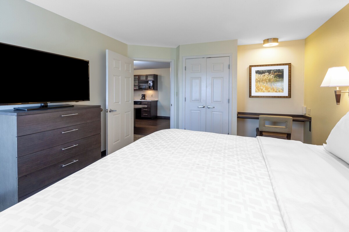 A hotel room with a bed, a TV, a desk with a lamp, a picture on the wall, and a view into the kitchen.