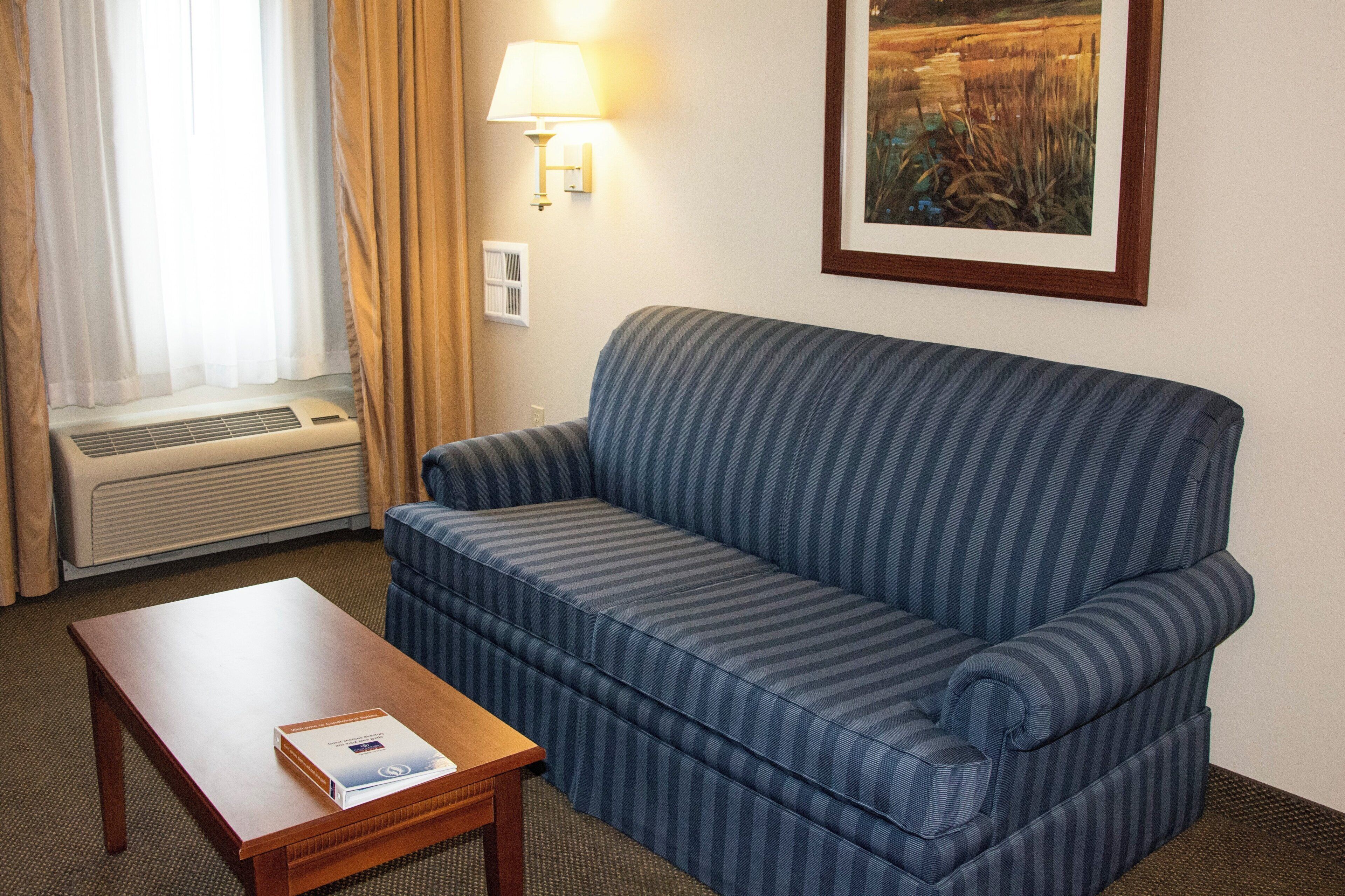 Una camera d'albergo con un divano a righe blu, un tavolino con un libro, una finestra con tende, una lampada a parete e un quadro incorniciato.