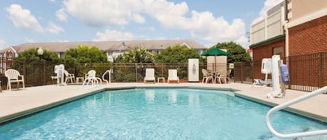 A rectangular swimming pool with clear blue water, surrounded by white lounge chairs and a brick building.