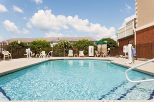 Una piscina rectangular con agua azul cristalina, rodeada de sillas de descanso blancas y un edificio de ladrillo.