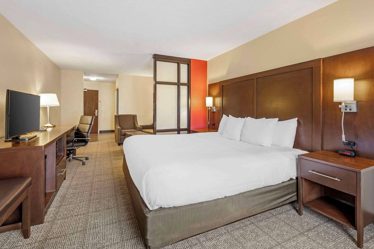 A hotel room with a large bed, a desk with a TV, a chair, a nightstand, and a lamp.