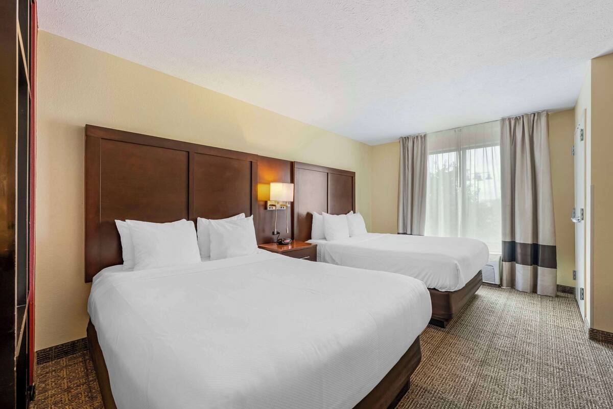 A hotel room with two beds, a wooden headboard, a nightstand with a lamp, and a large window with curtains.