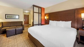 Een hotelkamer met een groot bed, een bank met bijpassende poufs, een nachtkastje met lamp en een ingelijste foto aan de muur.