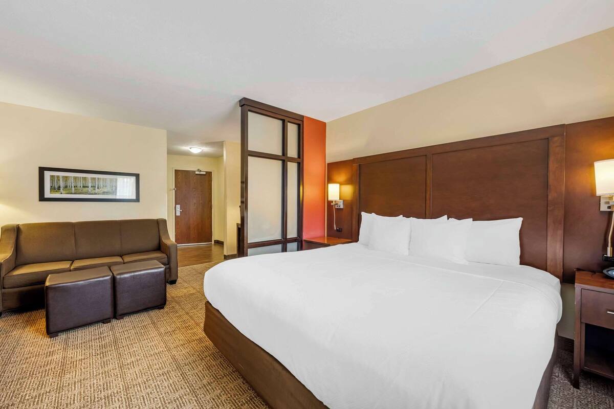 A hotel room with a large bed, a sofa with matching ottomans, a nightstand with a lamp, and a framed picture on the wall.