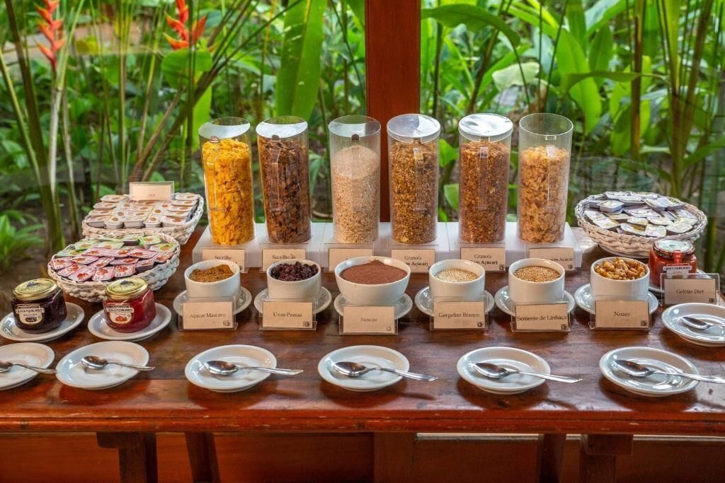 A buffet with various food items and condiments, including jars of preserves and bowls of grains and legumes, arranged on a wooden table with a view of greenery in the background.
