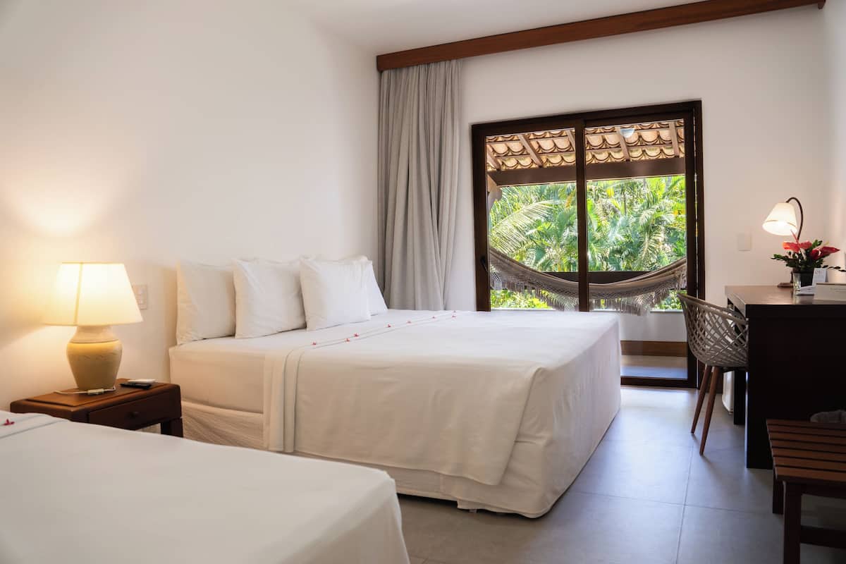 A hotel room with a bed, a desk, a chair, a lamp, and a view of greenery through a window.