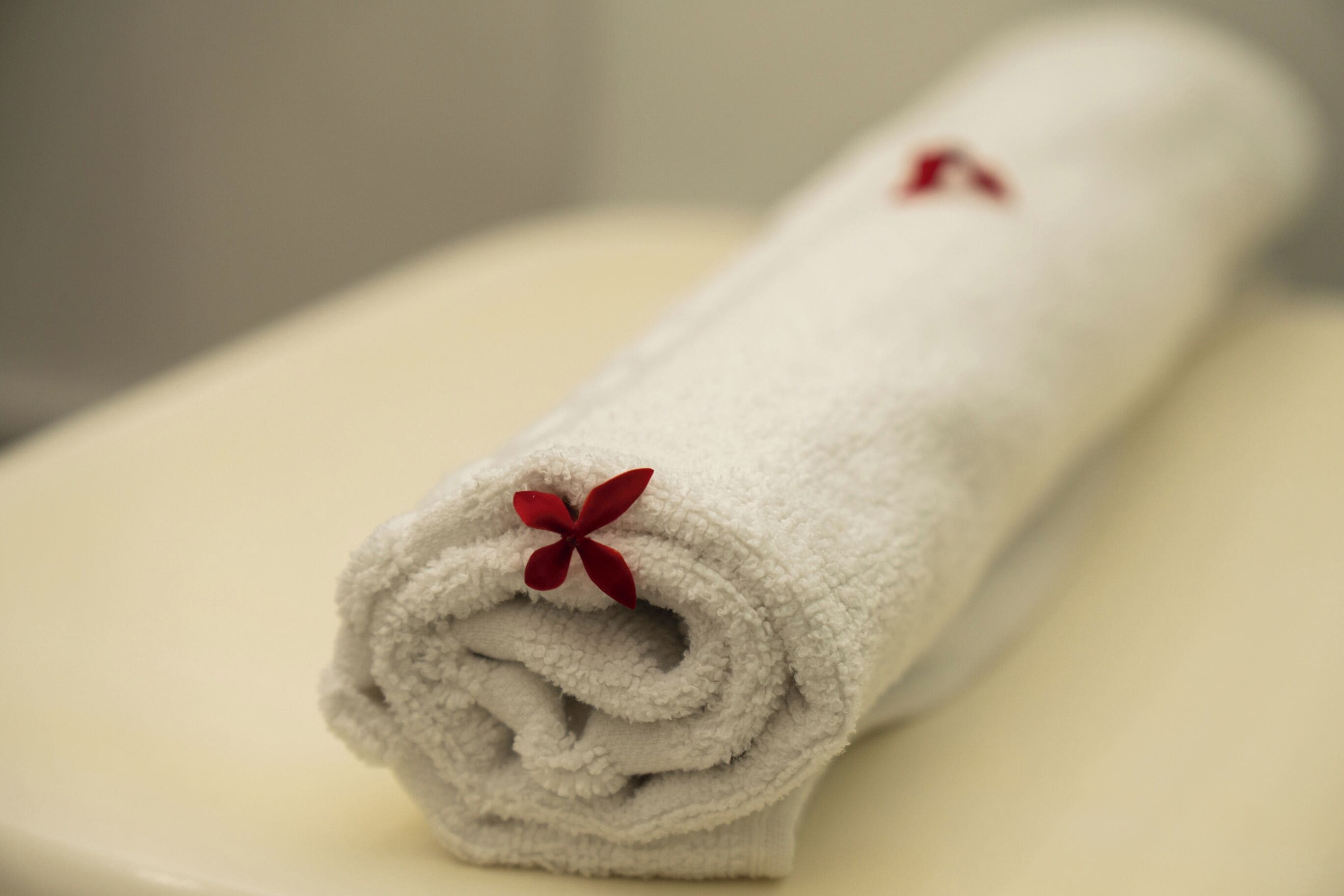 A rolled white towel with a red flower decoration on a white surface.