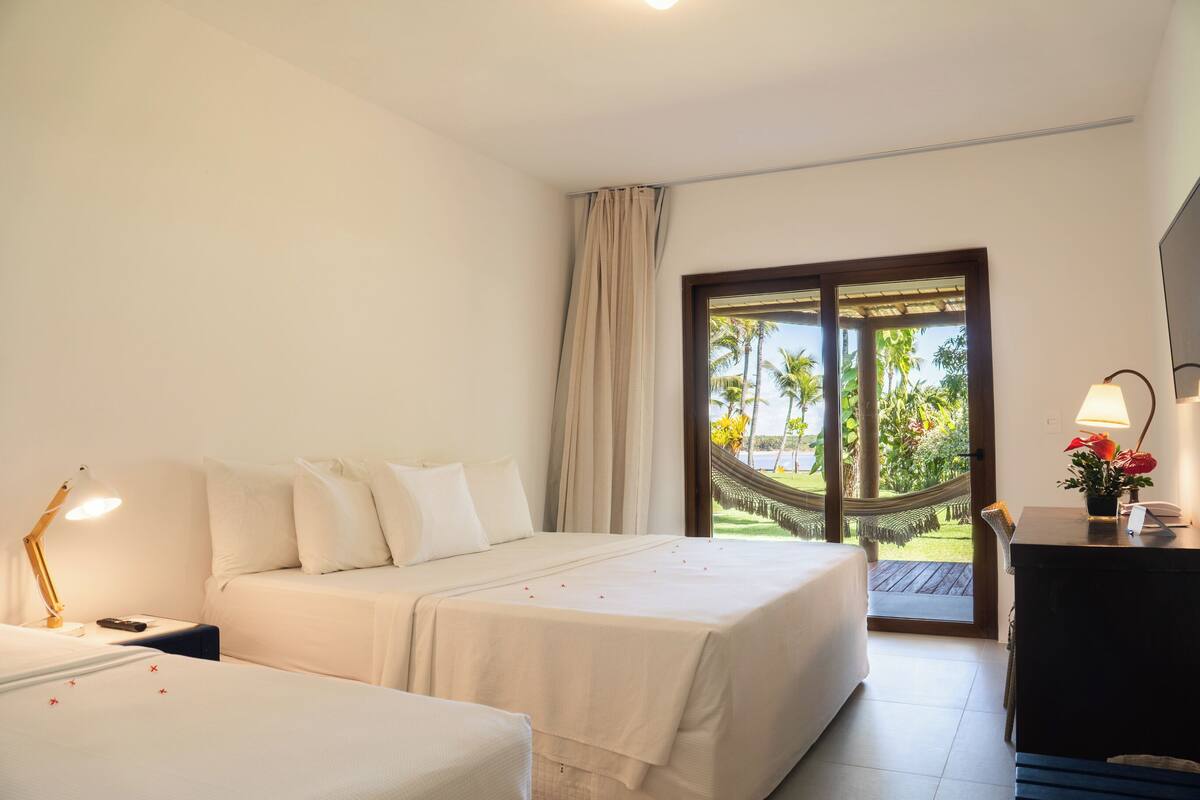 A hotel room with a bed, a desk, and a view of a hammock and greenery outside.