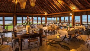 A restaurant with a thatched roof, large windows, and a view of the ocean. Tables are set with white tablecloths and wine glasses.