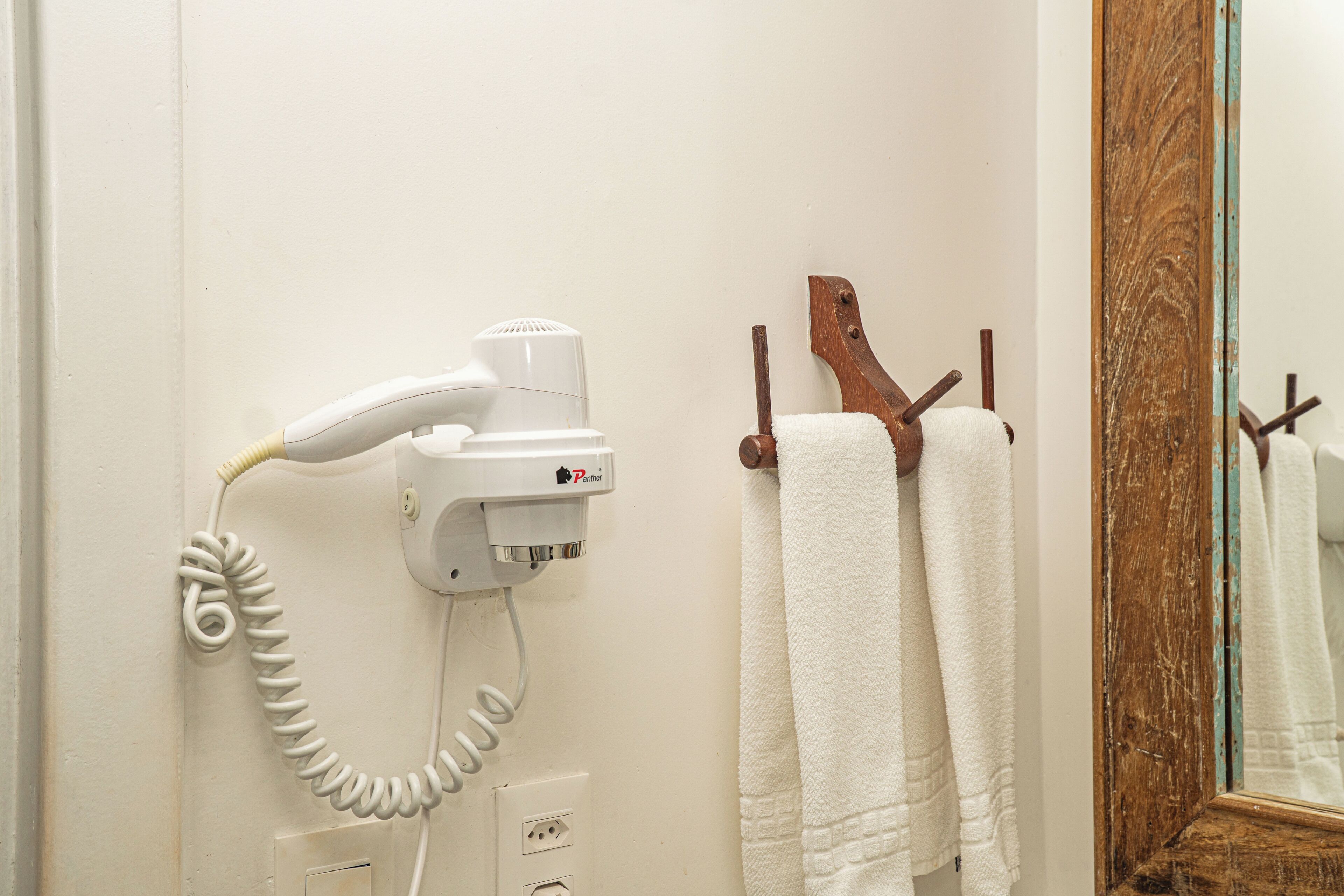 A hair dryer mounted on a wall, a wooden towel rack with two white towels, and a mirror with a wooden frame.
