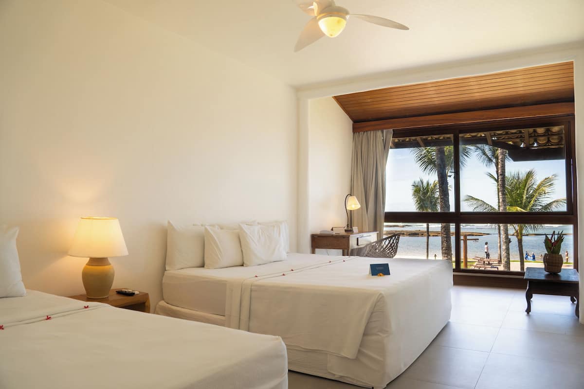 A hotel room with two beds, a ceiling fan, a nightstand with a lamp, a small table, and a view of the beach through large windows.