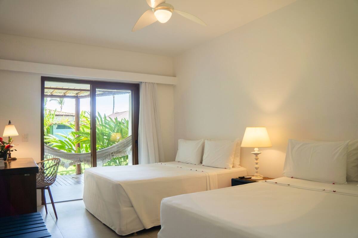 A hotel room with two beds, a desk, a chair, a hammock, and a view of greenery outside.