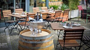Un área de comedor al aire libre con mesas y sillas de madera, un barril con un cubo de hielo y botellas, y una planta en maceta al fondo.