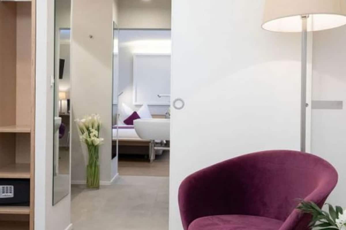 A modern hotel room with a purple armchair, a bedside table, and a lamp.
