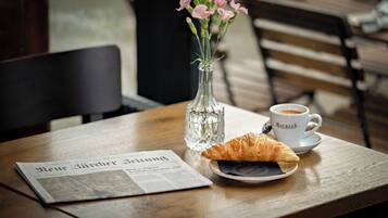 A newspaper, a croissant, a cup of coffee, and a vase with flowers on a wooden table.