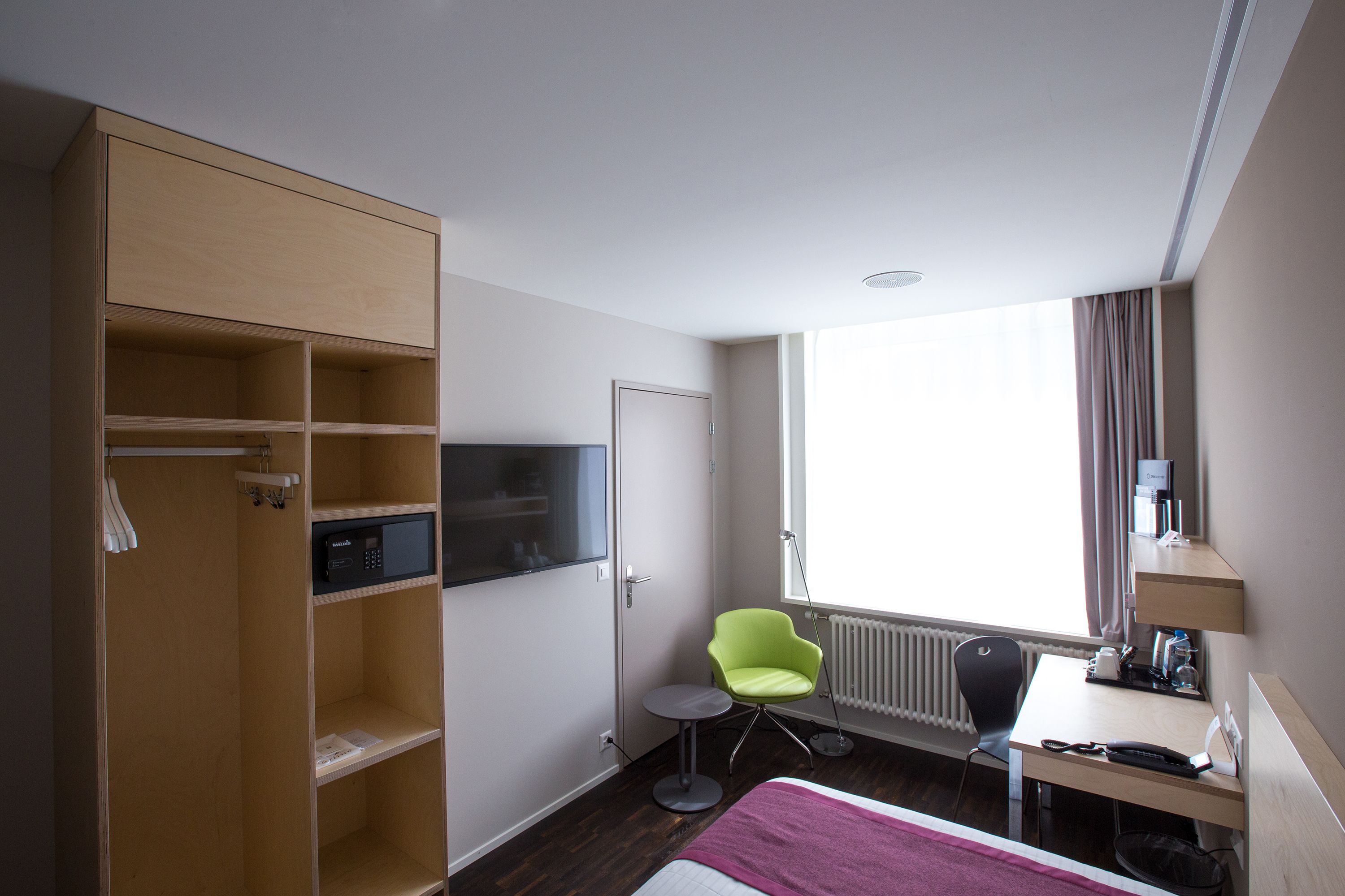 Habitació d'hotel amb llit, escriptori, cadira, armari, radiador i finestra.