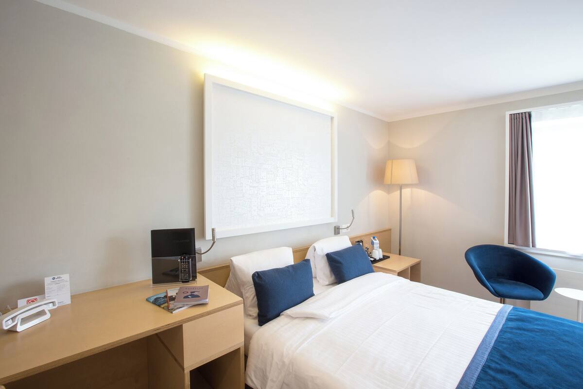 A hotel room with a bed, bedside tables, a desk, a chair, a lamp, and a window with curtains.