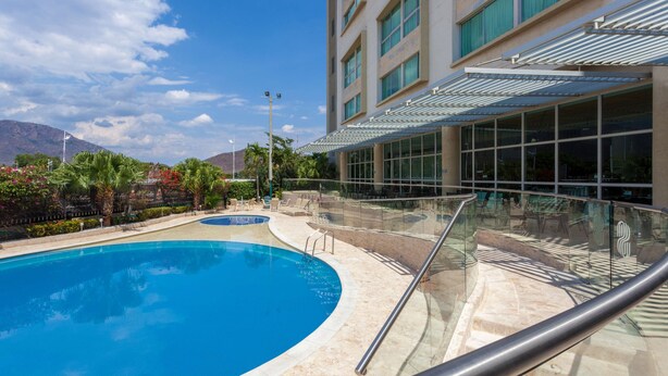 Una piscina con barandilla de vidrio, un edificio con grandes ventanales y palmeras al fondo.